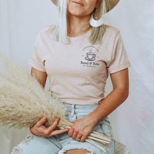 Pu&ograve; includere: T-shirt rosa chiaro con maniche arrotolate, con una grafica nera di una tazza da t&egrave; e piattino con il testo "Reed & Rite" e "St. Crispian's". La maglietta &egrave; abbinata a jeans azzurri.