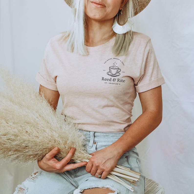 Pu&ograve; includere: T-shirt rosa chiaro con maniche arrotolate, con una grafica nera di una tazza da t&egrave; e piattino con il testo "Reed & Rite" e "St. Crispian's". La maglietta &egrave; abbinata a jeans azzurri.