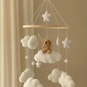 May include: A nursery mobile with a wooden frame, white clouds, stars, and a small teddy bear. The clouds and stars are made of soft, fluffy material. The mobile is suspended by white strings, creating a calming aesthetic.