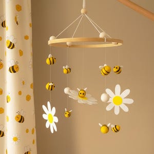 May include: A wooden baby mobile featuring yellow and black bees, white daisies, and white pom-poms suspended from a wooden ring. The mobile is hanging in front of a yellow and white patterned curtain.