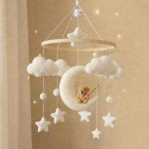 Può includere: Mobile per bambini con un anello di legno, nuvole bianche, stelle e una luna crescente. Un piccolo orsacchiotto è seduto sulla luna. Il mobile è sospeso da fili bianchi.