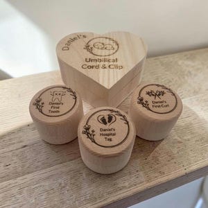 May include: A heart-shaped wooden box labelled Daniel's Umbilical Cord & Clip, accompanied by three small round wooden containers engraved with Daniel's First Tooth, Daniel's First Curl, and Daniel's Hospital Tag, each featuring floral wreath designs.
