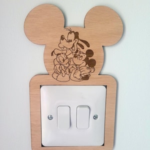 May include: Wooden light switch cover with Mickey Mouse, Goofy, and Donald Duck etched on it. The cover is shaped like Mickey Mouse's head and has a rectangular frame around the light switches. The light switches are white.