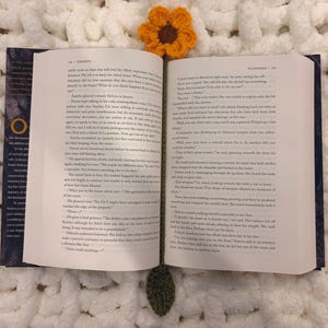 May include: An open hardcover book with printed text on white pages, featuring a crocheted bookmark with an orange flower top and a green leaf bottom, resting on a textured white surface.