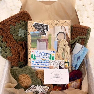 May include: A gift box containing a wrapped book with stickers, a crochet square, a crochet flower, a bookmark with a tassel, a small Twinings tea bag, and a card labeled Book N' Stitch. The book cover features text including I work to support my reading and Mystery.