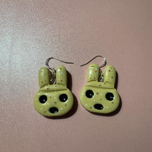 May include: A pair of yellow bunny-shaped earrings with black eyes and brown spots. The earrings are made of polymer clay and have silver hooks.