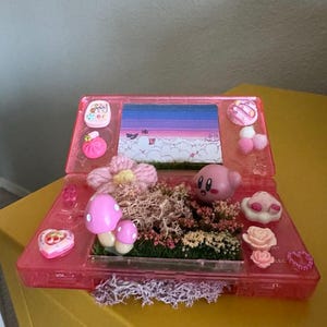 May include: A pink Nintendo DS handheld gaming console decorated with a miniature fairy garden. The garden features a pink Kirby character, mushrooms, flowers, and moss. The console's screen displays a pink and purple sky with clouds.