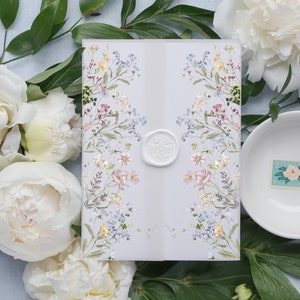 May include: A floral invitation with a white wax seal, surrounded by white peonies and greenery. The invitation features a delicate design of colorful flowers. A small white bowl with a floral stamp is also present.