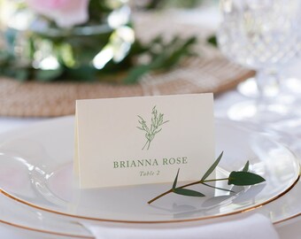 May include: A white place card with green foliage and the text "BRIANNA ROSE Table 2" on a white plate with a gold rim.