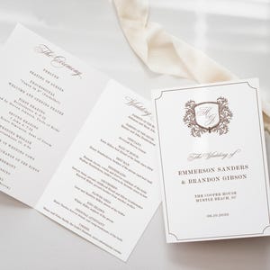 May include: Elegant wedding programs with a cream-colored background. The open program displays the ceremony details, while the cover features a decorative crest and the names of the couple, Emmerson Sanders and Brandon Gibson.