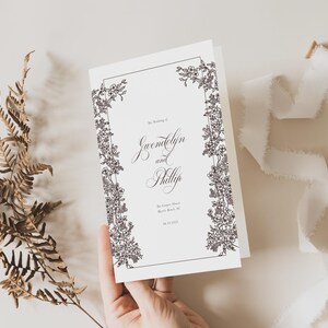 May include: A wedding invitation with floral border design. The invitation features the names "Gwendolyn and Phillip" in elegant script. A dried fern and white ribbon are also in the image. The invitation is on a white background.