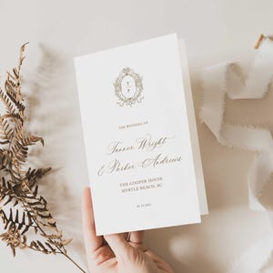 May include: A wedding program with the names "Tanner Wright & Parker Andrews" in elegant script. The program is cream-colored with a gold emblem and text. A sprig of dried foliage and a white ribbon are also in the image.