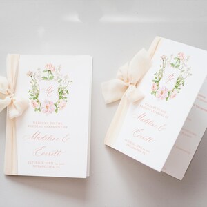 May include: Two wedding ceremony programs with floral designs and cream-coloured ribbons. The programs are white with pink text and a floral wreath design. The text reads "Welcome to the Wedding Ceremony of Madeline & Everett".