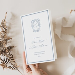 May include: A wedding program with a blue border and floral design. The program includes the names "Elaina Wright & Peter Andrick" and the wedding location. A white ribbon and dried fern are also in the image.
