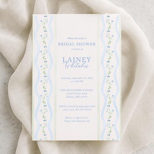 May include: A white invitation card with blue and green floral border details. The card reads "BRIDAL SHOWER" and includes event details. The invitation is on a white cloth background.