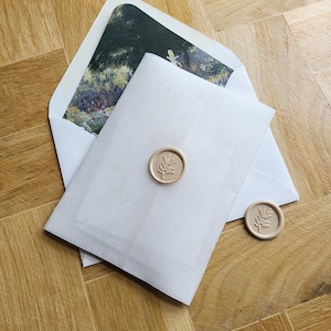 May include: A white envelope with a wax seal featuring a leaf design. The envelope is on a wooden surface.