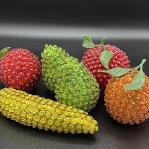 Pode incluir: Uma coleção de frutas decorativas, incluindo duas maçãs vermelhas, um pepino verde, uma laranja e uma espiga de milho amarela. Cada peça é coberta com pequenos enfeites redondos e brilhantes. A fruta tem folhas verdes.