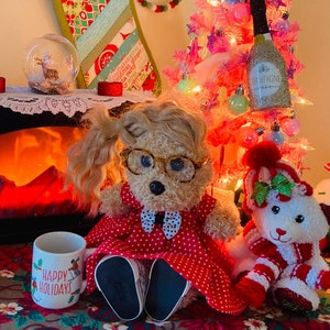 May include: A plush toy dog wearing glasses and a red dress with white polka dots sits in front of a fireplace with a mug that says "Happy Holidays". A pink miniature Christmas tree with lights and ornaments is in the background.