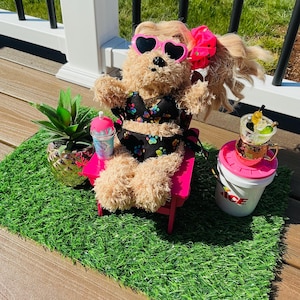 May include: A plush dog wearing heart-shaped sunglasses and a black bikini sits on a pink lounge chair. The dog has a pink bow in its hair and is surrounded by miniature accessories, including a drink and a potted plant, on a patch of green turf.