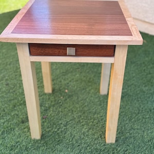 May include: A small wooden end table with a single drawer. The table top is made of a dark wood with a lighter wood border. The legs are made of a light wood. The table is sitting on a green grassy surface.