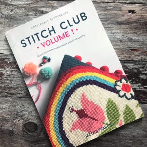 May include: A book titled "STITCH CLUB VOLUME 1" with a rainbow cross-stitch project visible. The book cover features the text "QUICK STITCH MODERN CROSS STITCH PROJECTS".  Also visible are colorful pom-poms and beads.