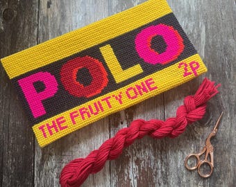 Peut inclure: Une pièce faite à la main, brodée au point de croix, avec le mot "POLO" en rose vif et rouge, sur un fond jaune et noir. Le texte "THE FRUITY ONE" est également visible. Une tresse de fil rouge et de petits ciseaux dorés sont à côté.