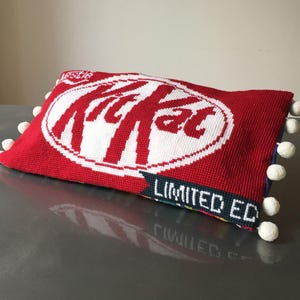 May include: Red and white knitted pillow with the word "KitKat" in a circle and the words "Limited Ed" on the bottom. The pillow has white pom-poms around the edges.