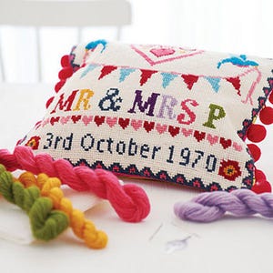 May include: A white pillow with colorful embroidery that reads "Mr. & Mrs. P 3rd October 1970". The pillow has a fringe border and pom-poms on the corners. There are colorful yarn skeins in the foreground.