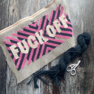 May include: Needlepoint canvas with the words "FUCK OFF" in white, set against a pink and navy blue starburst design. A skein of dark blue yarn and small scissors are next to the canvas on a wooden surface.