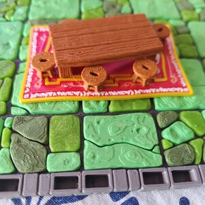 Op de afbeelding: Een miniatuur houten tafel en drie kleine krukjes op een roze en geel tapijt. De tafel en krukjes zijn bruin. Het tapijt ligt op een groen, getextureerd oppervlak dat lijkt op een stenen vloer.