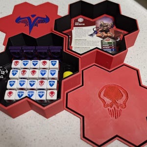 Puede incluir: Organizador de juegos hexagonal rojo y negro con dados, piezas de juego y cartas. El organizador presenta un emblema de calavera y un logotipo azul y rojo. Incluye un dado de 20 caras en blanco y negro y varios dados blancos con símbolos de calavera roja y cresta azul.