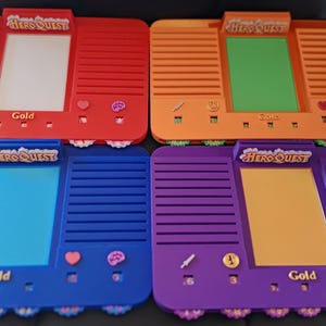 May include: Four brightly coloured HeroQuest game boards in red, orange, blue, and purple. Each board features a rectangular screen area and decorative elements such as hearts, brains, and a sword. The word "Gold" is printed on each board.