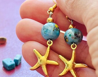 Puede incluir: Un par de pendientes de estrella de mar dorados con cuentas azules y doradas. Los pendientes tienen un estilo de gancho de pez. Los pendientes son un accesorio con temática de playa, sostenidos por una mano.