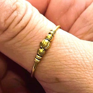 Może przedstawiać: Złoty pierścionek z serią małych, okrągłych koralików i większym, teksturowanym koralikiem pośrodku. Pierścionek ma cienką, delikatną obrączkę. Pierścionek jest noszony na palcu.