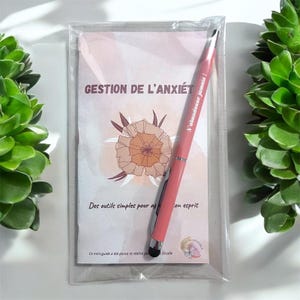 Peut inclure: Un livret rose et blanc avec le titre "Gestion de l'anxiété" et un stylo rose avec le texte "N'abandonne jamais!" imprimé dessus. Le livret a un motif floral et le texte "Des outils simples pour affronter son esprit".