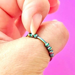 Peut inclure: Une bague anti-stress de couleur arc-en-ciel avec de petites perles irisées. La bague est composée d'une fine bande métallique incurvée avec plusieurs petites perles colorées qui peuvent être déplacées le long de la bande. La bague est présentée sur un doigt sur fond rose.