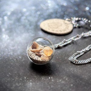 Puede incluir: Un collar de cadena de plata con un colgante de esfera de vidrio que contiene arena y conchas marinas. La esfera es transparente y muestra la arena y las conchas en su interior.