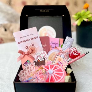 Könnte beinhalten: Eine schwarze Geschenkbox gefüllt mit rosa und weißen Artikeln, darunter ein Heft mit dem Titel "Gestion de l'anxiété", ein Aufkleber mit der Aufschrift "Blame it on the A-A-A-A", ein rosa-weißer Fidget Spinner, ein rosa Lutscher und ein rosa Stift.