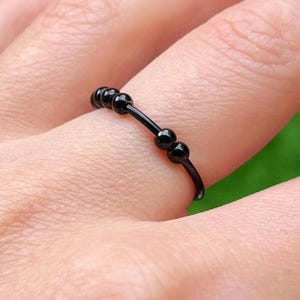 Op de afbeelding: Een zwarte ring met kleine, ronde kralen. De ring wordt om een vinger gedragen en de kralen zijn gelijkmatig verdeeld over de band. De ring lijkt van metaal te zijn en heeft een eenvoudig, elegant ontwerp.