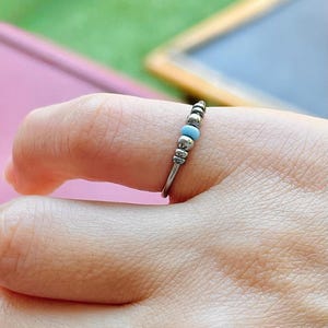 Puede incluir: Un anillo plateado con un diseño de cuentas. Presenta pequeñas cuentas plateadas y dos cuentas de color azul claro. El anillo se lleva en un dedo, destacando su estilo delicado y minimalista. Es una pieza de joyería.