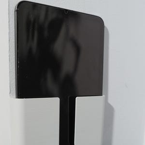 Op de afbeelding: Een witte wandhouder voor tablets met een gladde, matte afwerking en een rechthoekig zakontwerp. De houder is bevestigd aan een plat oppervlak en bevat een zwarte tablet met een reflecterend scherm, verticaal geplaatst voor weergave.