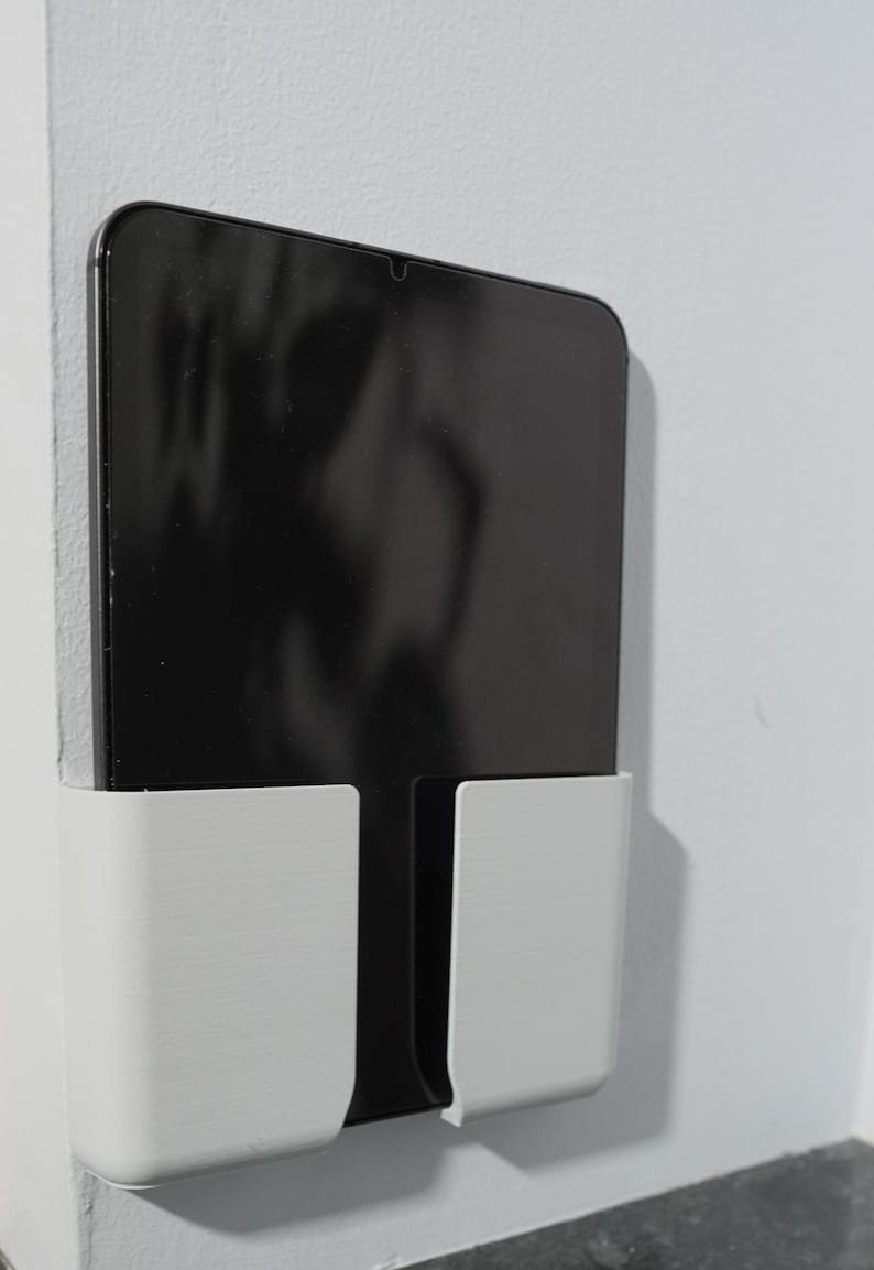 Op de afbeelding: Een witte wandhouder voor tablets met een gladde, matte afwerking en een rechthoekig zakontwerp. De houder is bevestigd aan een plat oppervlak en bevat een zwarte tablet met een reflecterend scherm, verticaal geplaatst voor weergave.