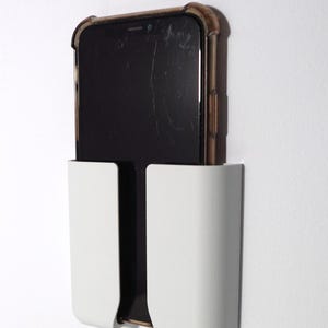 Op de afbeelding: Een witte rechthoekige telefoonhouder voor aan de muur met een U-vormige uitsparing in het midden. Een zwarte smartphone met een donker doorschijnend beschermhoesje is verticaal in de houder geplaatst en rust stevig in het witte kunststof frame.