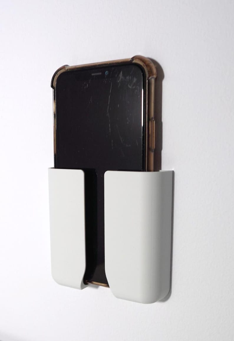 Op de afbeelding: Een witte rechthoekige telefoonhouder voor aan de muur met een U-vormige uitsparing in het midden. Een zwarte smartphone met een donker doorschijnend beschermhoesje is verticaal in de houder geplaatst en rust stevig in het witte kunststof frame.