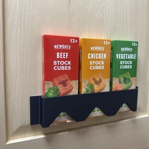 Op de afbeelding: Drie dozen Newgate bouillonblokjes in rood, geel en groen, met de labels "Beef", "Chicken" en "Vegetable". Elke doos heeft een afbeelding van de smaak en de tekst "12x". De dozen staan in een zwarte houder.
