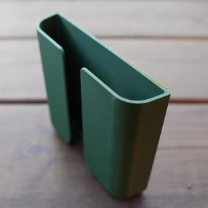 Op de afbeelding: Een donkergroene, U-vormige kunststof houder met een gladde, matte afwerking. Het object heeft twee parallelle verticale compartimenten met open bovenkanten en een plat achterpaneel, met een dunne gele plakstrip langs de bovenrand voor montage.