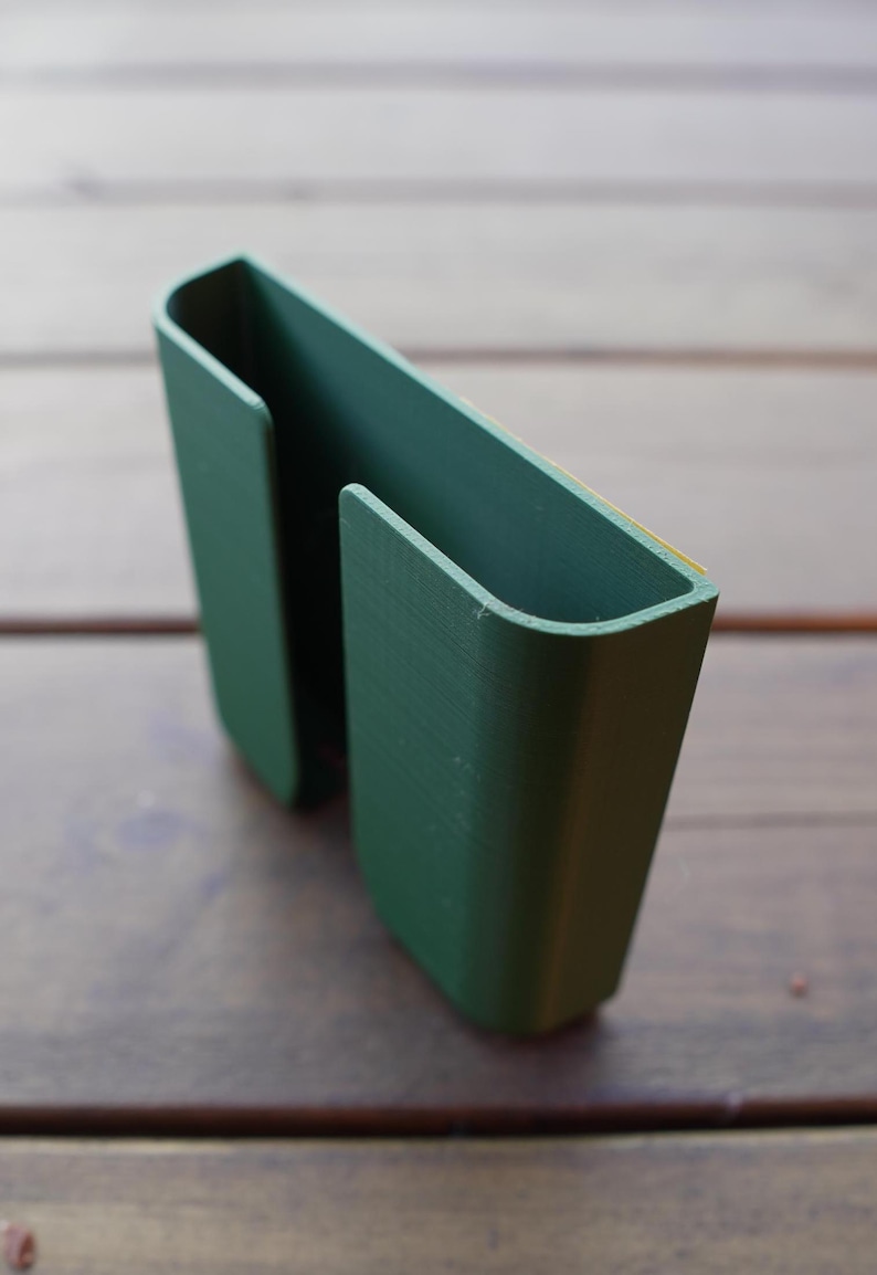 Op de afbeelding: Een donkergroene, U-vormige kunststof houder met een gladde, matte afwerking. Het object heeft twee parallelle verticale compartimenten met open bovenkanten en een plat achterpaneel, met een dunne gele plakstrip langs de bovenrand voor montage.