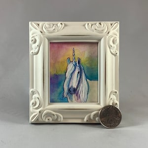 May include: A white framed watercolor painting of a unicorn with a rainbow background. The unicorn is facing forward with its head tilted slightly to the side. The painting is in a white frame with ornate details.