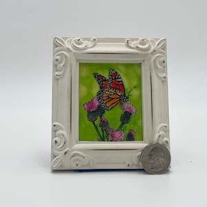 Può includere: Un dipinto in miniatura incorniciato in bianco di una farfalla monarca su sfondo verde con fiori rosa. La cornice ha una finitura bianca invecchiata e dettagli decorativi.