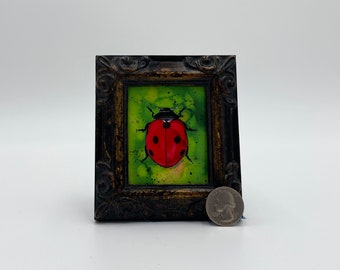 May include: A framed painting of a red ladybug with black spots on a green background.